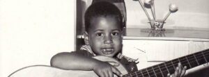 Foto em preto e branco de 1975 mostrando Geovane Souza ainda criança, um menino negro de cabelos curtos e crespos. Ele segura um violão acústico maior que seu corpo e sorri com inocência para a câmera, usando uma camisa de gola listrada. A imagem ilustra a pureza da infância antes da consciência do preconceito racial e estético.