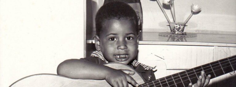 Foto em preto e branco de 1975 mostrando Geovane Souza ainda criança, um menino negro de cabelos curtos e crespos. Ele segura um violão acústico maior que seu corpo e sorri com inocência para a câmera, usando uma camisa de gola listrada. A imagem ilustra a pureza da infância antes da consciência do preconceito racial e estético.
