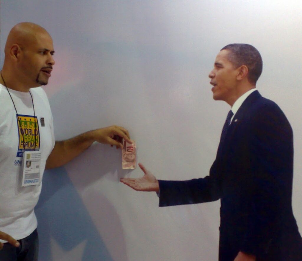 Geovane Souza handing a 10 reais note to a cardboard cutout of Barack Obama with his hand outstretched.