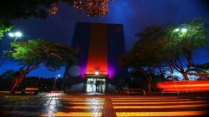Vista noturna do Paço Municipal de São José dos Campos iluminado, com trilhas de luz de veículos em movimento no primeiro plano.