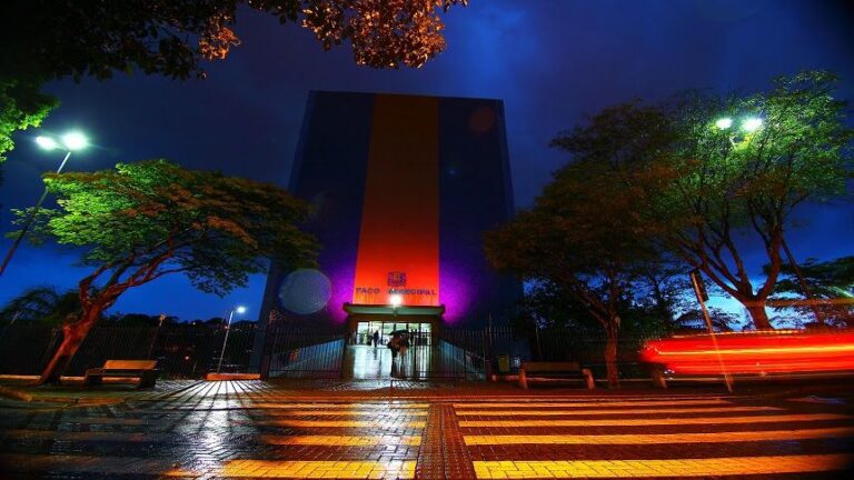 Vista noturna do Paço Municipal de São José dos Campos iluminado, com trilhas de luz de veículos em movimento no primeiro plano.