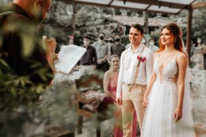 Bride and groom Maria Luiza (redhead) and Eduardo (in suspenders) smiling emotionally at the altar of Solar das Palmeiras, listening to a personalized reading of their story by celebrant Geovane Souza (highlighted on the left).