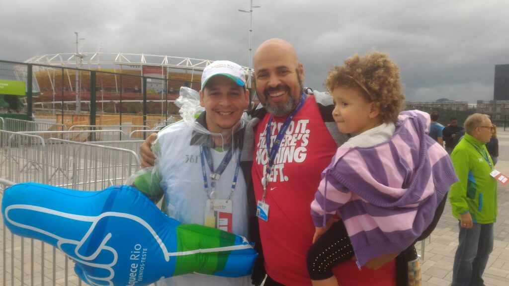 Exterior photo at the Olympic Park. On the left, Affonso Junior smiles wearing a raincoat and holding an inflatable "hand" from the Aquece Rio campaign. On the right, the author Geovane Souza, in a red shirt, smiles while hugging his daughter, Gabriela Cristina, who is wearing a purple striped jacket and looking to the side. In the background, the exterior structure of an Olympic arena can be seen.