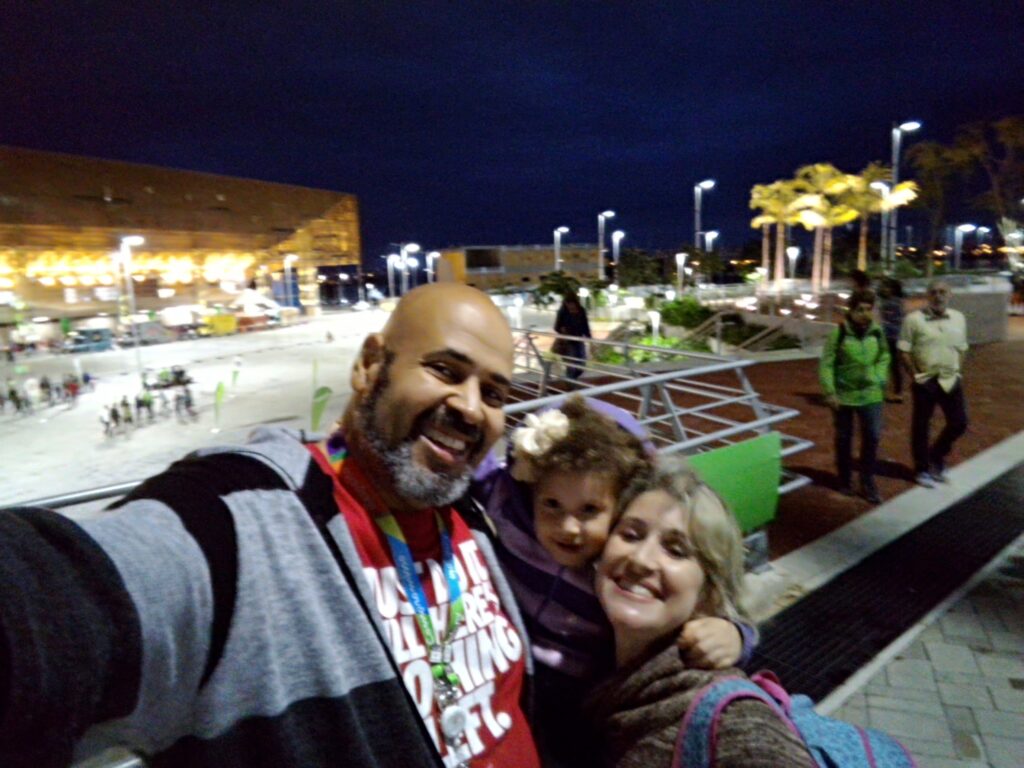 Night selfie of Geovane Souza, his wife Carina, and daughter Gabriela Cristina at the Barra Olympic Park. They smile, embracing in the foreground. In the background, the facade of the Arena of the Future shines intensely with its yellow and gold night lighting.