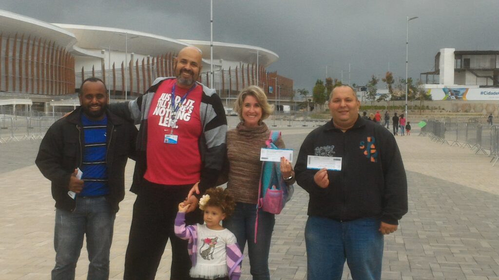 Group photo at the Barra Olympic Park on a cloudy day. From left to right: Ítalo smiles holding a ticket; the author Geovane Souza (the 2.07m tall giant) wears a red Nike shirt and holds his daughter Gabriela Cristina's hand; his wife, Carina, displays a ticket next to Maurício, who also shows his invitation. In the background, the Aquatic Stadium and the park's walkway can be seen.