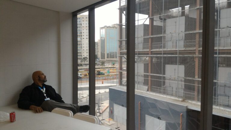 Representing the acclimatization period at the MPC (Main Press Center) of Rio 2016: photo by the author, Geovane Souza, sitting in an office at Avenida Abelardo Bueno, nº 4801, in Barra da Tijuca. He is relaxed in a chair, looking out through a large glass window. The view shows the construction of the neighboring building and modern residential buildings in the background.