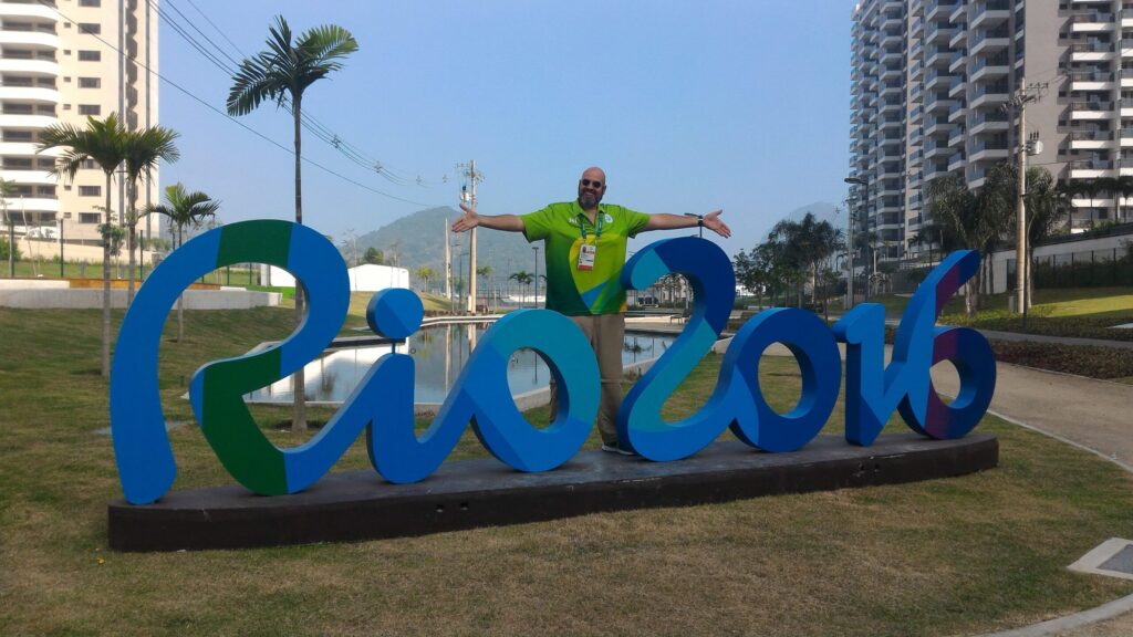 Foto de Geovane Souza, vestindo a camisa verde de Líder EVS, de braços abertos diante da escultura do logo Rio 2016. A foto foi tirada dentro da Vila Olímpica e Paralímpica, uma área de segurança máxima e acesso restrito aos atletas e credenciados. Ao fundo, veem-se os prédios residenciais dos atletas.